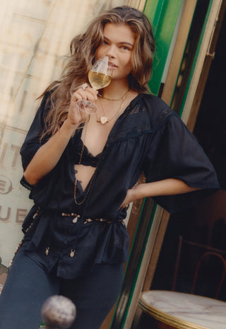 a-woman-with-long-wavy-brown-hair-wearing-a-black-bohemian-style-sheer-blouse-and-matching-pants-holding-a-glass-of-white-wine-in-front-of-her-face-she-is-posed-in-front-of-a-rustic-doorway
