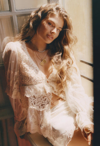 a-woman-with-long-wavy-light-brown-hair-wearing-a-sheer-cream-lace-lingerie-set-with-a-matching-long-sleeve-robe-posed-in-warm-sunlight-near-a-window